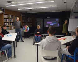 Wieczór quizowy w bibliotece. Bibliotekarz stoi obok kolorowego koła fortuny i patrzy na uczestników siedzących przy białych stołach. W tle dwie wolontariuszki przy flipcharcie oraz ekran z napisem „Wieczór Quizowy”. Na środku sali, tyłem do obiektywu, siedzi kobieta na wózku