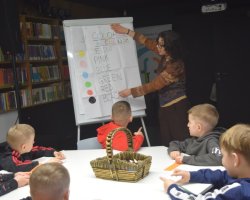 Grupa chłopców siedzi przy białym stole, słuchając wolontariuszki. Kobieta stoi przy tablicy flipchart, na której widnieje napis „COLORS IN ENGLISH” oraz lista nazw kolorów z odpowiadającymi im kolorowymi kropkami. Na stole stoi wiklinowy koszyk z kredkami