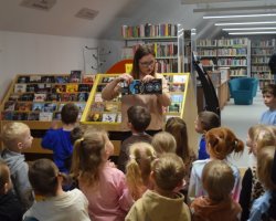 Bibliotekarka w okularach stoi przed grupą małych dzieci w dziale zbiorów audiowizualnych. Kobieta pokazuje dzieciom otwarte opakowanie z płytą CD, tłumacząc zasady wypożyczania multimediów. Dzieci z zaciekawieniem patrzą na prezentację