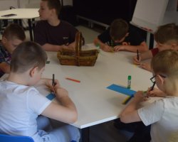 Zbliżenie na grupę sześciu chłopców siedzących wokół białego stołu i zaangażowanych w rysowanie. Na blacie leżą kredki, kleje w sztyfcie oraz paski kolorowego papieru. Na środku stołu stoi podłużny, wiklinowy koszyczek z przyborami plastycznymi