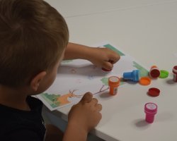 Widok zza ramienia małego chłopca siedzącego przy białym stole, który stawia kolorowe pieczątki na karcie pracy z ilustracją jelenia. Po prawej stronie leżą porozrzucane plastikowe stempelki oraz ich zatyczki